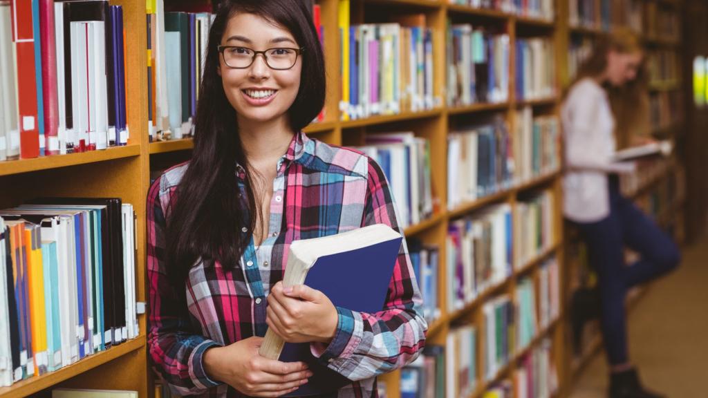 Smiling student leaning against bookshelves holding book at the university
