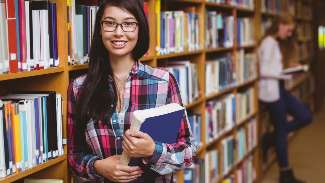 Smiling student leaning against bookshelves holding book at the university