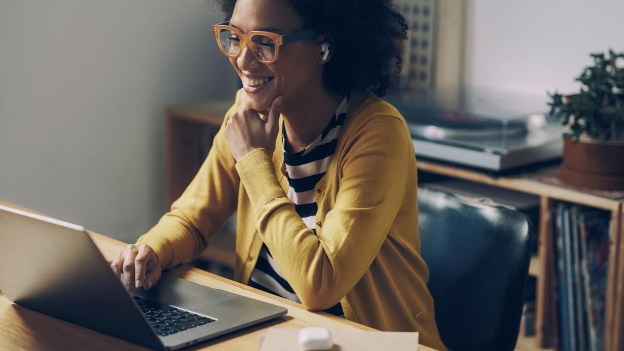 Smiling African American Woman Wearing Glasses and Wireless Earphones Makes a Video Call on her Laptop Computer at her Home Office