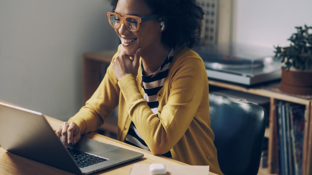 Smiling African American Woman Wearing Glasses and Wireless Earphones Makes a Video Call on her Laptop Computer at her Home Office
