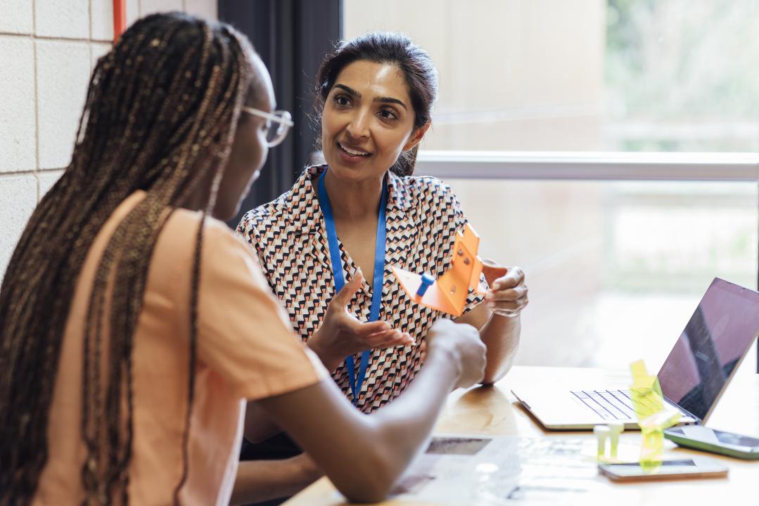 A shot of a secondary school teacher sitting with a student, discussing their Design Technology work, in a classroom within a school in Gateshead, England.